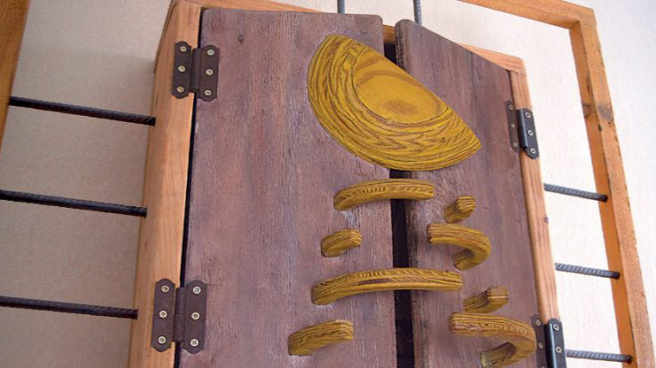 Informania IV, a mixed-media artwork made from recycled materials: two hinged wooden panels with carved, yellow-painted elements representing a satellite, mounted against a wall with visible metal rods, recycled industrial wood, texture, and symbolism.