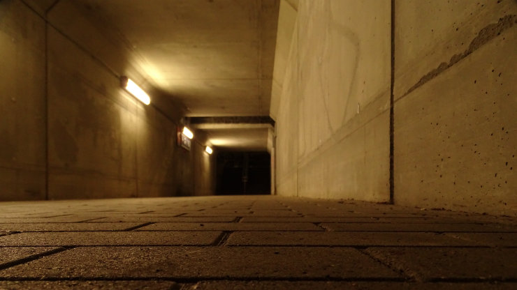 Liminal tunnel vision, a low-angle photograph of an empty concrete tunnel with tiled flooring and warm wall lights, receding into darkness and evoking solitude, transition, and quiet unease.